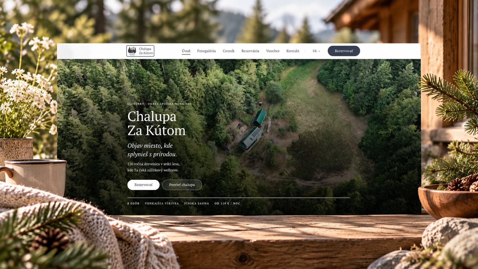 Úvodní stránka webu Chalupa Za Kútom s leteckým záběrem lesa na obrazovce na dřevěném stole na verandě, vedle hrnek, deka a větvičky jehličí