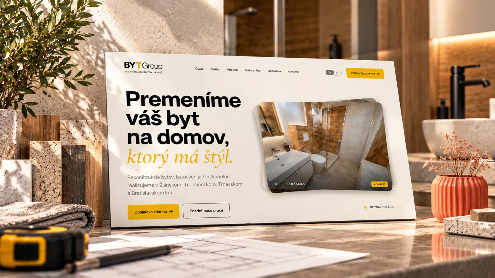 Úvodní stránka webu BYTGroup na obrazovce mezi vzorky kamene a dřeva, metrem, výkresem a korálovou vázou.